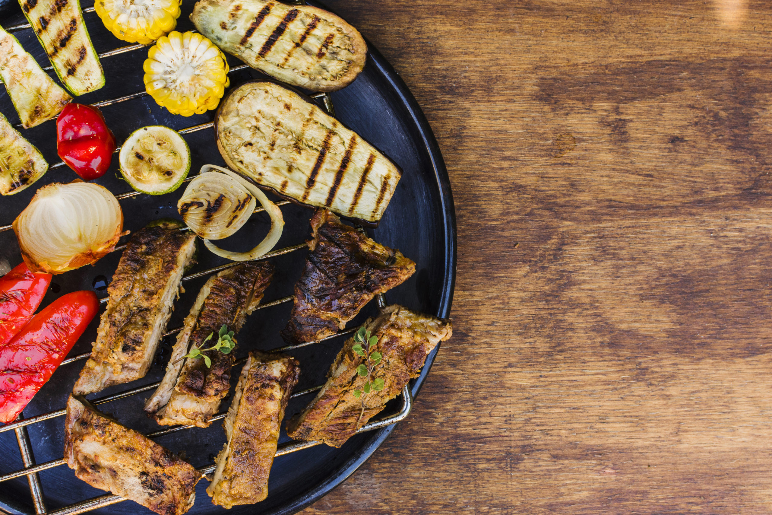 Foto con vista cenital de una mesa de madera con un plato negro a la izquierda de la imagen con verduras y carne a la brasa